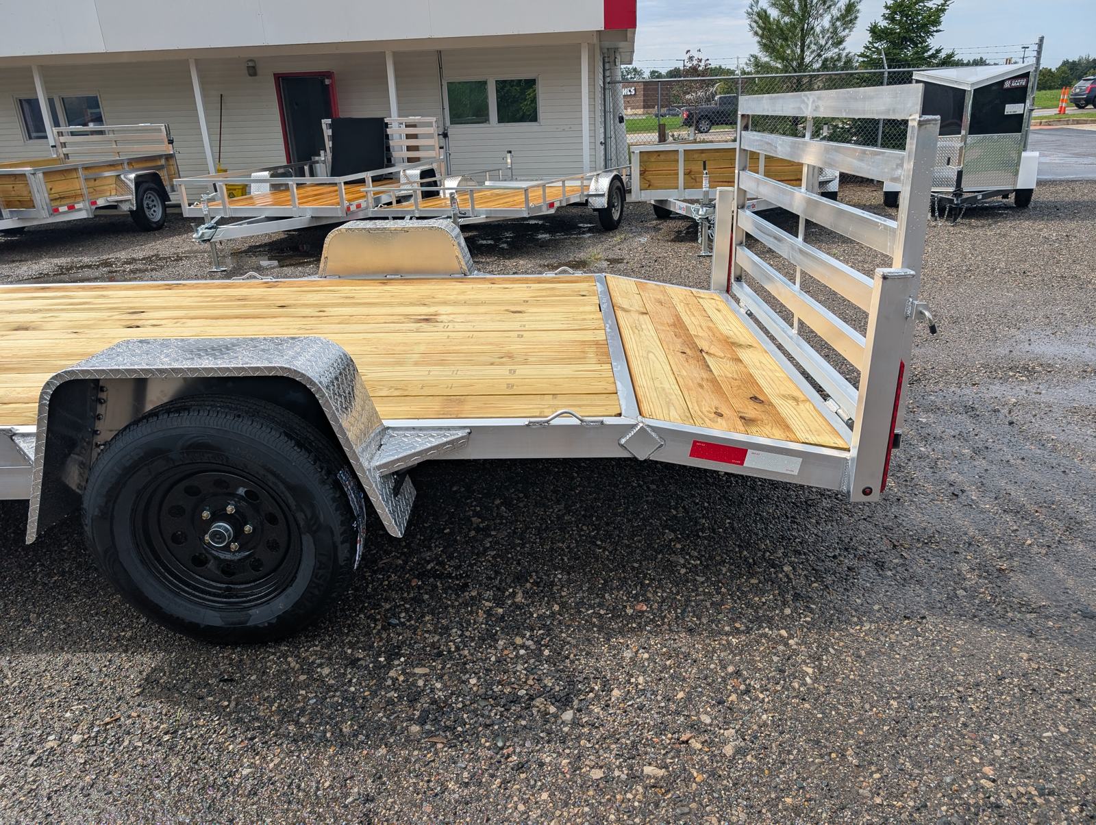 Close-up of the rear corner of the Silver Ox 7x14 Oxtail Sport showing the aluminum diamond-plate fender, wheel, tail light, and the gate hinge and latch with the slatted gate standing upright