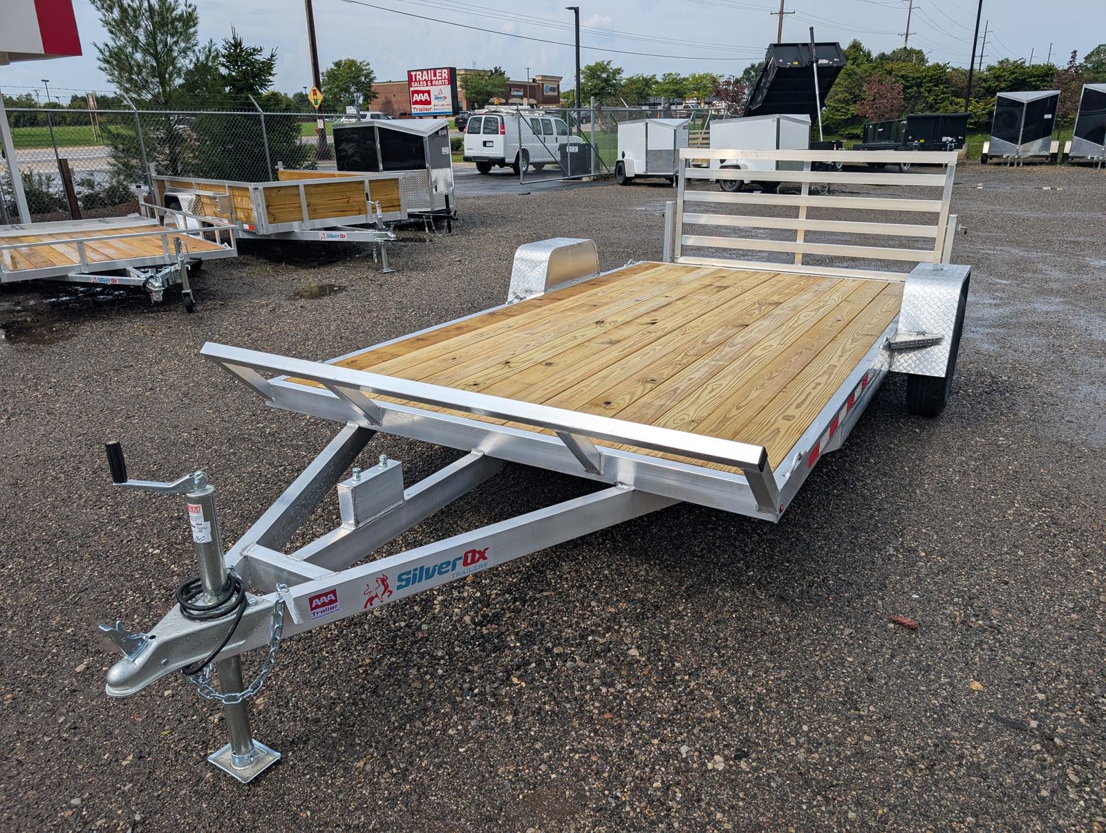Front three-quarter view of the Silver Ox 7x14 Oxtail Sport showing the aluminum A-frame tongue, coupler, tongue jack, and red Silver Ox and AAA Trailer decals, with the open deck and dovetail rear in the background
